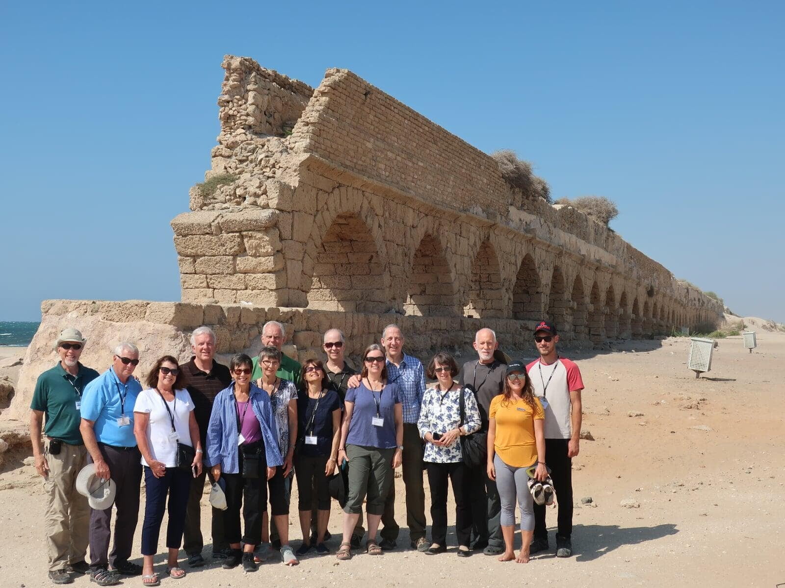 A group of tourists posing in front of an ancient aqueduct built by Herod the Great at Caesarea Maritima, Israel, on a clear day.